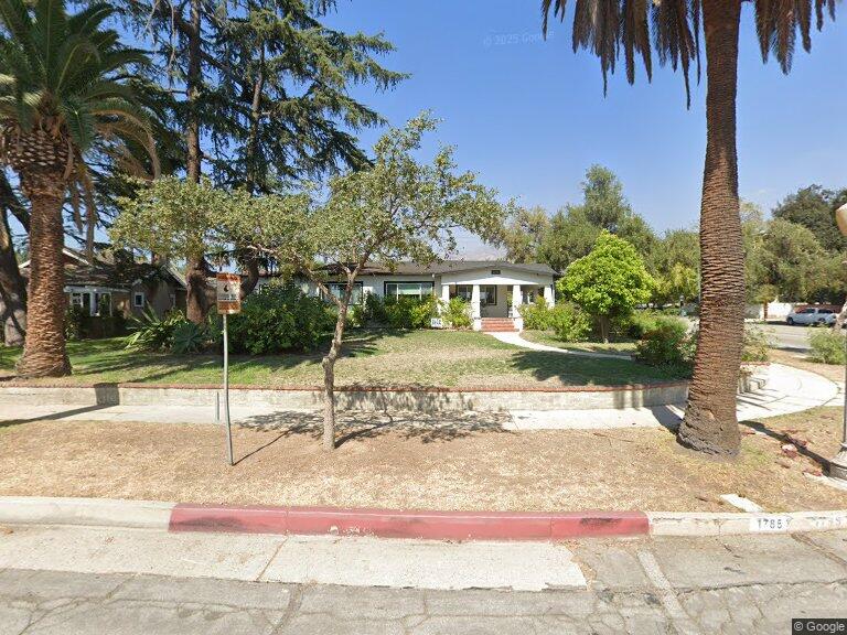 Quaint house with green lawn, trees, and palm-lined street under clear sky