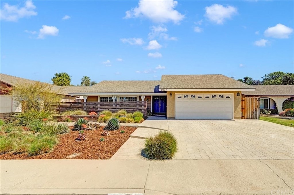 Single-story house with beige walls, a two-car garage, blue front door, and minimalist desert landscaping. The sky is clear with few clouds.