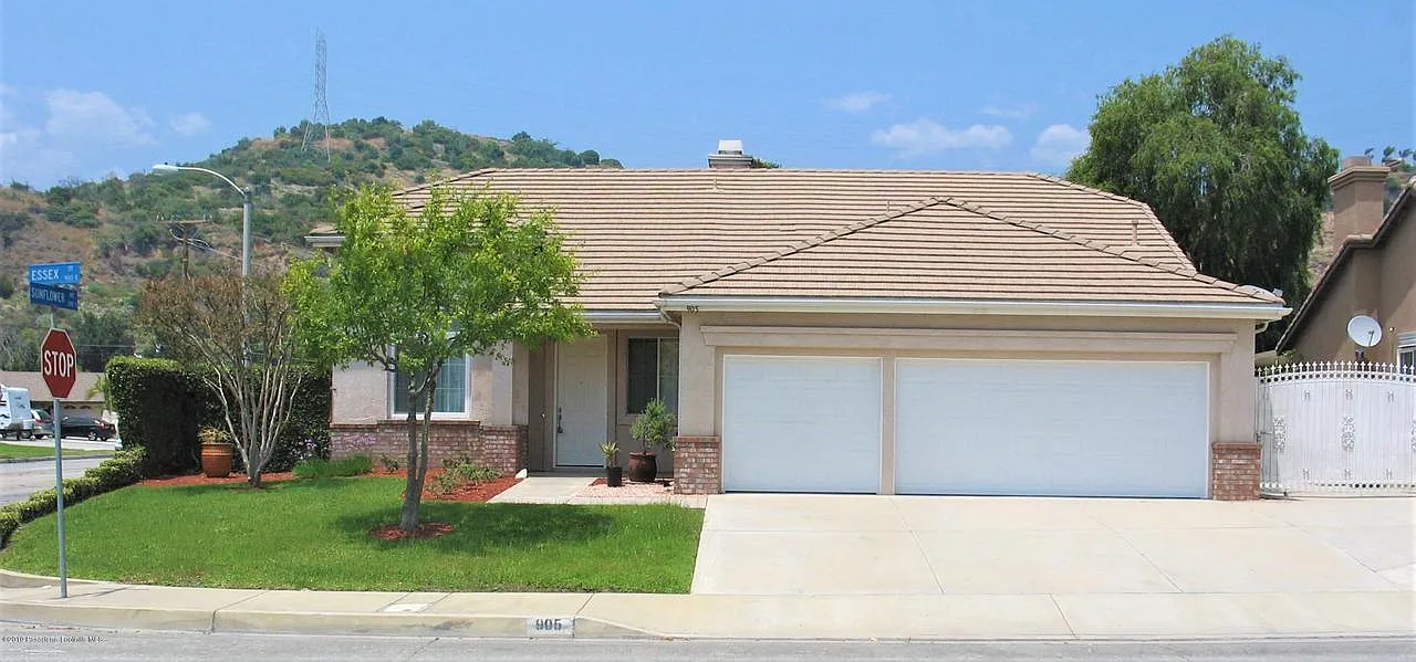 A suburban house with a beige exterior, two-car garage, and a brown tile roof. It’s on a corner lot with a manicured lawn, a tree, and potted plants.