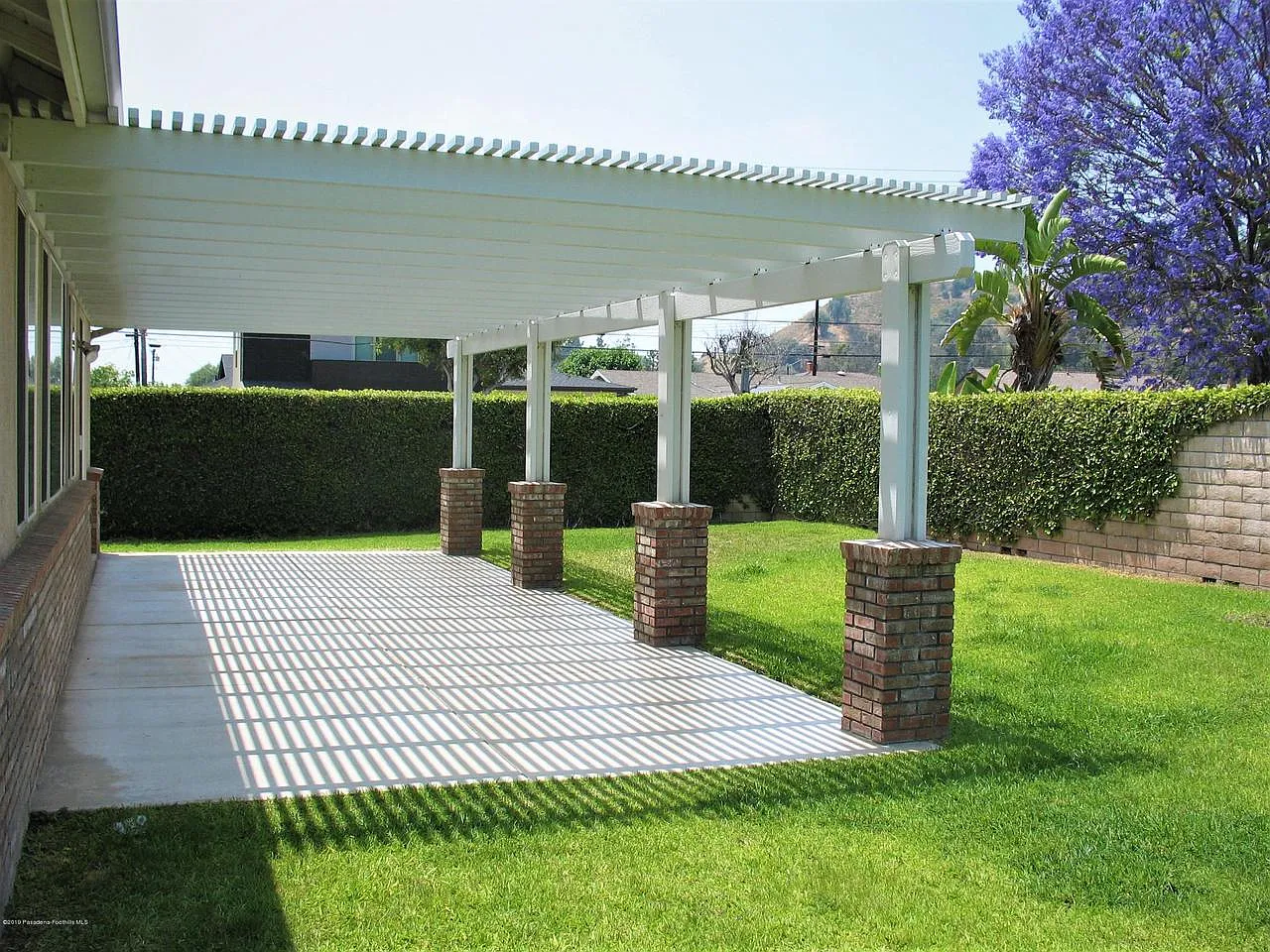 Patio with white pergola, striped shadows, brick pillars, lawn, and flowering tree