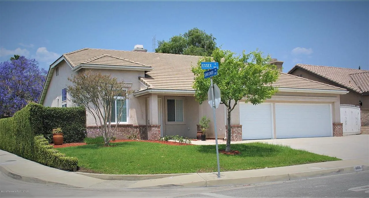 Corner suburban house with beige siding, tan roof, lawn, trees, and street signs