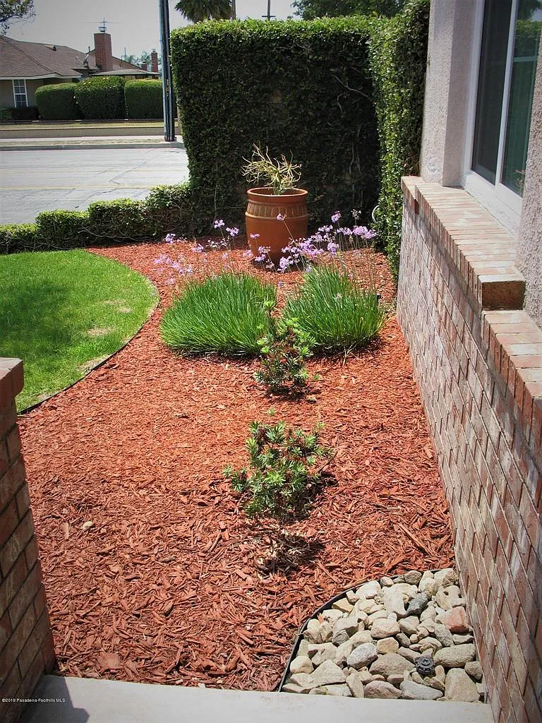 Narrow garden with red mulch, stone border, shrubs, flowers, and terracotta pot