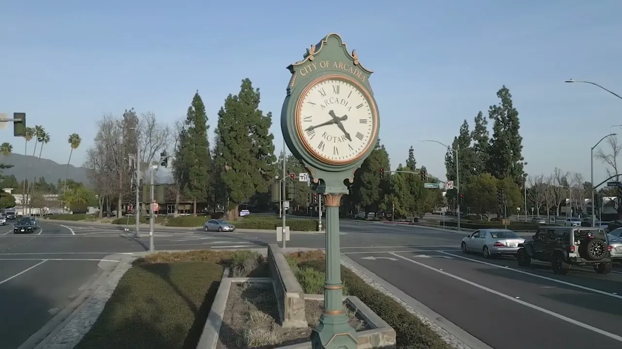 A large clock on a pedestal in a roundabout, surrounded by trees and cars.