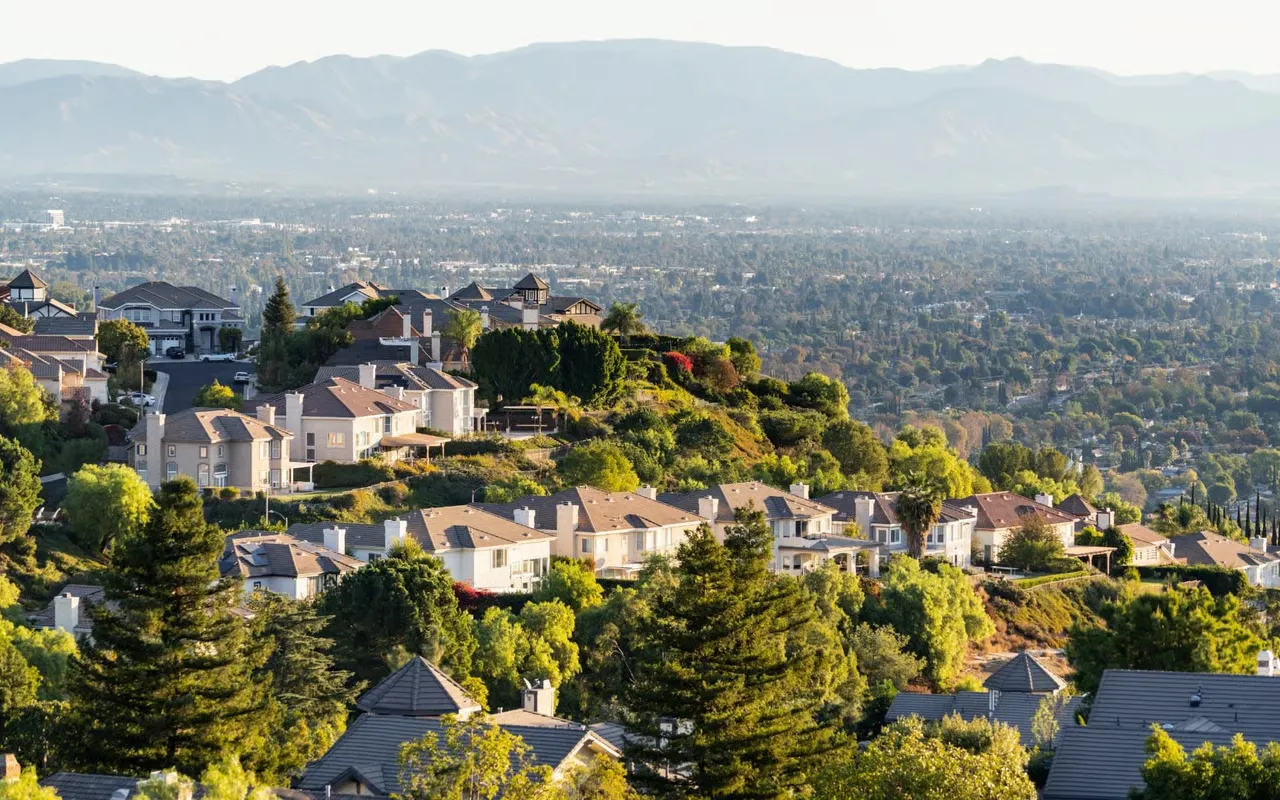Aerial view of a suburban neighborhood with houses on hills, surrounded by trees and mountains in the distance.